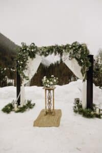 Ein Hochzeitsaltar in einer verschneiten Landschaft mit einem hölzernen Bogen, der mit weißem Stoff behangen und mit Grünpflanzen und Blumen geschmückt ist. Unter dem Bogen steht ein kleiner Holztisch mit einem Blumenarrangement auf einem von Grünpflanzen gesäumten Weg.