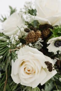 Ein Strauß mit weißen Rosen, Tannenzweigen, kleinen weißen Blumen und braunen Tannenzapfen vor einem unscharfen Hintergrund. Das Arrangement hebt eine Mischung aus Texturen und natürlichen Elementen hervor.