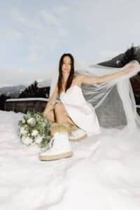 Eine Frau in einem kurzen, schulterfreien weißen Kleid und Schleier sitzt im Schnee. Sie hält einen Strauß weißer Blumen mit Grün in der Hand. Sie trägt pelzbesetzte weiße Stiefel und posiert vor einer Kulisse aus schneebedeckten Bergen und einem teilweise bewölkten Himmel.