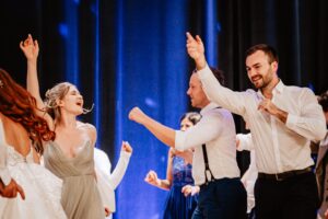 Menschen tanzen fröhlich bei einer Indoor-Veranstaltung. Die Gruppe ist formell gekleidet, die Männer tragen weiße Hemden und die Frauen elegante Kleider. Im Hintergrund leuchtet ein blaues Licht, das eine festliche Atmosphäre schafft.
