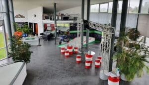 Das Bild zeigt eine großzügige Indoor-Anlage mit einem Fahrsimulator. Es gibt rot-weiße Absperrungen, Pflanzen und einen Empfangstresen. Mehrere Bildschirme und Banner mit der Aufschrift „Rasch Group“ sind sichtbar, und rundherum sind Sitzbereiche angeordnet.