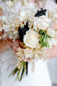 Eine Braut hält vor dem Hintergrund eines weißen Brautkleides einen Strauß mit weißen Rosen, schwarzen Blumen und zarten weißen Blüten, durchsetzt mit grünen Blättern und braunen Zweigen.