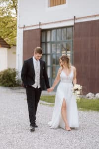 Braut und Bräutigam gehen Hand in Hand auf einem Kiesweg. Der Bräutigam trägt einen schwarzen Anzug, die Braut ein weißes Kleid mit Schlitz und hält einen Blumenstrauß in der Hand. Sie stehen im Freien in der Nähe eines Gebäudes mit großen Fenstern und rostfarbenen Paneelen.