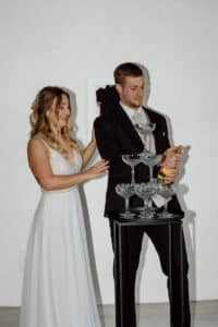 Braut und Bräutigam sind auf einer Hochzeit und gießen gerade Champagner in einen Turm aus Gläsern. Der Bräutigam hält die Flasche bereit zum Einschenken. Die Braut, ein weißes Kleid tragend, lächelt und legt ihre Hand auf den Arm des Bräutigams. Im Hintergrund ist eine schlichte weiße Wand.
