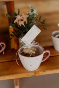 In einer rosa-weißen Tasse mit Herzmuster liegt ein kleines, mit einer Schleife verschnürtes Geschenk mit der Aufschrift „Emma“. Im Hintergrund ist auf einer Holzoberfläche ein verschwommenes Blumenarrangement zu sehen.