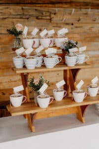 Auf einem Holzregal stehen mehrere Keramikbecher mit Etiketten, die jeweils mit kleinen Leckereien oder Geschenken gefüllt sind. Kleine Vasen mit rosa Rosen und Grünpflanzen schmücken die Regale. Im Hintergrund befinden sich rustikale Holzwände.