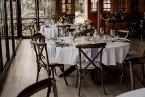 Elegant gedeckte Esstische mit weißen Tischdecken und Holzstühlen sind mit floralen Tischdekorationen und ordentlich angeordnetem Geschirr geschmückt und befinden sich in einem sonnendurchfluteten Raum mit großen Fenstern und Holzdekor.