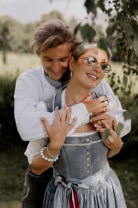 Ein Paar umarmt sich im Freien. Ein Mann umarmt eine Frau von hinten. Sie lächelt, trägt eine Sonnenbrille und ein traditionelles Kleid. Beide tragen Ringe und Armbänder. Die Szene ist üppig und grün und lässt einen sonnigen Tag in der Natur erahnen.