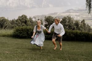 Ein Paar in traditioneller Kleidung läuft fröhlich barfuß über eine Wiese. Im Hintergrund ist eine malerische Aussicht auf Berge und einen See unter einem dunstigen Himmel zu sehen. Die Frau trägt ein Kleid, der Mann ein Hemd und Shorts.