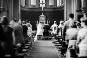 Ein Brautpaar geht den Mittelgang einer Kirche entlang, während die Gäste stehen und zuschauen; das Bild ist schwarz-weiß mit einer leichten Unschärfe an den Rändern und konzentriert sich auf das Paar, das sich dem Altar nähert.