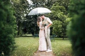 Ein Paar steht eng aneinander gedrängt unter einem durchsichtigen Sonnenschirm in einem grünen Park. Die Frau hält einen Blumenstrauß in der Hand. Umgeben von Bäumen und Grün blicken sie sich an einem bewölkten Tag liebevoll an.