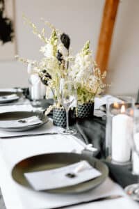 Elegante Tischdekoration mit schwarzen Tellern, einem Weinglas und weißen Blumen auf einer weißen Tischdecke. Schwarze Servietten und Kerzenhalter ergänzen das Arrangement und schaffen ein elegantes Ambiente.