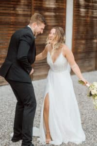 Braut und Bräutigam verbringen einen fröhlichen Moment draußen. Die Braut, in einem weißen Kleid mit hohem Schlitz, hält einen Blumenstrauß und lacht mit dem Bräutigam, der einen schwarzen Anzug trägt. Sie stehen auf Kies, im Hintergrund eine Holzmauer.