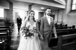 Eine Braut im weißen Kleid mit einem Blumenstrauß und ein Bräutigam im Anzug gehen lächelnd Hand in Hand den Mittelgang einer Kirche entlang. Im Hintergrund sind Kirchenbänke und eine weitere Person zu sehen, die hinter ihnen geht. Das Bild ist schwarz-weiß.