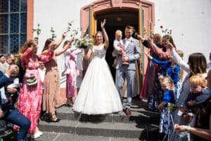 Braut und Bräutigam verlassen mit einem kleinen Kind im Arm ein Gebäude, während die Gäste jubeln und Rosen hochhalten. Das Paar lächelt und winkt, umgeben von gut gekleideten Freunden und Familienmitgliedern, die an einem sonnigen Tag im Freien feiern.