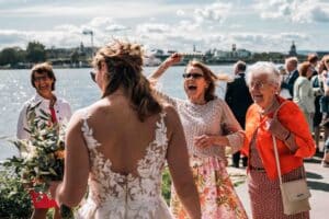 Eine Braut in einem weißen Spitzenkleid begrüßt an einem sonnigen Tag lächelnde Frauen am Wasser. Eine Frau jubelt mit erhobenem Arm, während eine andere, in Rot, neben ihr lacht. Im Hintergrund mischen sich weitere Gäste.