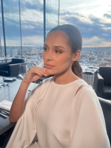 Eine Frau in einem weißen Kleid sitzt an einem Tisch in einem modernen, gehobenen Restaurant mit großen Glasfenstern und Blick auf die Stadtlandschaft unter einem teilweise bewölkten Himmel. Sie stützt ihr Kinn auf ihre Hand und blickt nachdenklich zur Seite.