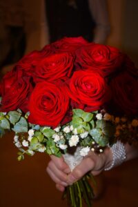 Eine Person hält einen Strauß leuchtend roter Rosen, akzentuiert mit kleinen weißen Blüten und grünem Laub. Der Strauß ist elegant in weiße Spitze gewickelt.