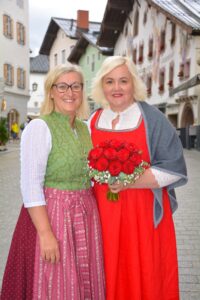 Zwei Frauen in traditioneller bayerischer Kleidung stehen auf einer Kopfsteinpflasterstraße. Eine hält einen Strauß roter Rosen in der Hand. Im Hintergrund sind bemalte Gebäude unter einem bewölkten Himmel zu sehen.