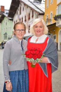 Zwei Frauen stehen zusammen in einer malerischen Straße mit farbenfrohen Häusern. Eine hält einen Strauß roter Rosen in der Hand und trägt ein rot-weißes Kleid mit einem grauen Schal; die andere trägt ein blaues Kleid und eine graue Jacke. Beide lächeln.