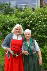 Zwei Frauen stehen zusammen in einem Garten und lächeln in die Kamera. Die Frau links hält einen Strauß roter Rosen und trägt ein rotes Kleid mit einem grauen Schal. Die Frau rechts trägt ein grünes Kleid mit einer weißen Bluse. Im Hintergrund ist grünes Laub zu sehen.