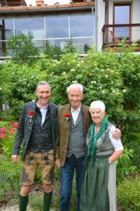Drei Menschen posieren glücklich in einem Garten mit üppigem Grün und Blumen. Ein älterer Mann und eine ältere Frau, beide traditionell gekleidet, stehen lächelnd neben einem jüngeren Mann in ähnlicher Kleidung. Im Hintergrund ist ein Gebäude mit großen Fenstern zu sehen.