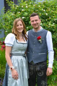 Ein lächelndes Paar steht in traditioneller bayerischer Kleidung in einem Garten. Die Frau trägt ein hellblaues Dirndl, der Mann eine graue Weste mit Stickerei und Lederhosen. Im Hintergrund sind weiße Blumen zu sehen.