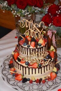 Eine zweistöckige Hochzeitstorte, dekoriert mit frischen Erdbeeren, Blaubeeren und Brombeeren, beträufelt mit Schokolade. Auf den Holzaufsätzen stehen die Aufschriften „Mr & Mrs“ und „Just Married“. Im Hintergrund sind rote Rosen und Grünpflanzen zu sehen.