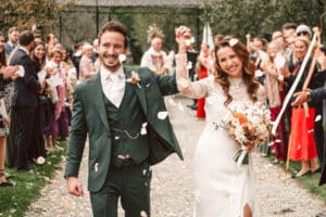 Ein fröhliches Brautpaar schreitet händchenhaltend den Gang entlang, umgeben von jubelnden Gästen. Die Braut trägt ein weißes Kleid und hält einen Blumenstrauß in der Hand, während der Bräutigam einen grünen Anzug trägt. Im Freien werden sie mit Blütenblättern überschüttet.
