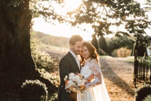Braut und Bräutigam stehen dicht beieinander, beleuchtet vom Sonnenlicht, das durch die Bäume fällt. Sie hält einen Strauß weißer und rosa Blumen in der Hand, und beide tragen formelle Kleidung. Der Weg und das Laub schaffen eine ruhige, romantische Atmosphäre.
