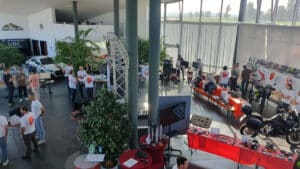 Eine geschäftige Indoor-Tech-Expo mit regen Austausch unter den Besuchern. Einige Besucher tragen passende T-Shirts. Verschiedene technische Exponate, darunter Motorradteile und Bildschirme, sind auf Tischen aufgebaut. Große Fenster lassen Tageslicht herein. Grünpflanzen stehen überall.