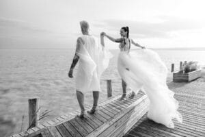 Ein elegant gekleidetes Paar spaziert händchenhaltend über einen Holzsteg am Meer. Die Frau trägt ein wallendes Kleid, beide sind tätowiert. Die Szene ist heiter und bietet einen ruhigen, weiten Blick auf das Wasser unter einem sanften Himmel.
