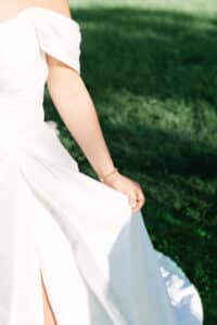 Eine Person in einem weißen, schulterfreien Kleid steht auf grünem Gras und hält das Kleid mit einer Hand. Sonnenlicht beleuchtet ihren Arm und ihr Kleid, und an ihrem Handgelenk ist ein zartes Armband zu sehen.