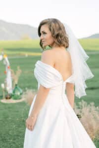 Eine Braut in einem schulterfreien weißen Brautkleid und Schleier steht draußen auf grünem Gras und blickt über ihre Schulter. Im Hintergrund sind sanfte Berge und verschwommene Hochzeitsdekorationen zu sehen.