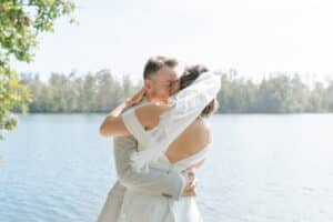 Braut und Bräutigam umarmen und küssen sich draußen an einem ruhigen See mit Bäumen im Hintergrund. Die Braut trägt ein weißes Kleid und einen Schleier; der Bräutigam einen hellen Anzug. Sonnenlicht schafft eine sanfte, romantische Atmosphäre.