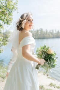 Eine lächelnde Braut in einem schulterfreien weißen Kleid und Schleier hält einen grünen Blumenstrauß, während sie an einem sonnigen Tag im Freien an einem See steht.
