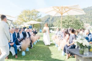 Eine Braut in einem weißen Kleid schreitet durch einen Gang im Freien, der von Gästen gesäumt ist, die zu beiden Seiten sitzen. Es ist sonnig, große Sonnenschirme spenden Schatten, im Hintergrund sind Berge zu sehen. Getrocknete Blumenarrangements schmücken den Gang.