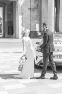 Eine Braut in einem weißen Kleid hält einen Blumenstrauß in der Hand und lächelt zu ihrem Bräutigam im dunklen Anzug auf. Sie stehen draußen neben einem weißen Auto und Steinsäulen und teilen einen freudigen Moment.