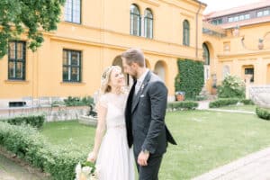 Braut und Bräutigam lächeln sich in einem grünen Innenhof mit einem gelben Gebäude im Hintergrund an. Die Braut hält einen Blumenstrauß in der Hand und trägt ein weißes Kleid; der Bräutigam trägt einen dunklen Anzug.