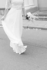 Eine Braut in einem langen weißen Kleid mit einem Blumenstrauß in der Hand geht auf einer Straße. Das Foto ist schwarz-weiß, ihr Gesicht ist nicht zu sehen.
