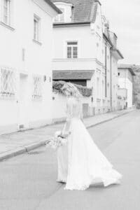 Eine Braut in einem langen, fließenden weißen Kleid und einem Spitzentop geht mit einem Blumenstrauß in der Hand eine ruhige Straße entlang, im Hintergrund sind elegante weiße Gebäude zu sehen. Das Bild ist in Schwarzweiß.