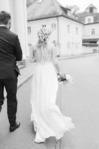Eine Braut in einem langen weißen Kleid und einem Spitzentop geht mit einem Blumenstrauß in der Hand neben einem Mann im dunklen Anzug eine ruhige Straße entlang. Das Foto ist schwarz-weiß und beide sind von hinten zu sehen.