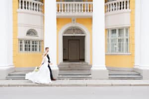 Braut und Bräutigam gehen Hand in Hand an einem großen gelben Gebäude mit weißen Säulen und Bogenfenstern vorbei, das Kleid der Braut weht hinter ihr her.
