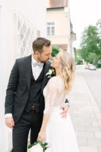 Braut und Bräutigam stehen dicht beieinander auf einem Bürgersteig und lächeln sich liebevoll an. Die Braut trägt ein weißes Kleid mit Spitzenärmeln und hält einen Blumenstrauß in der Hand, während der Bräutigam einen dunklen Anzug mit Fliege trägt.