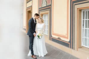 Braut und Bräutigam stehen dicht beieinander, lächeln und halten einen Blumenstrauß in der Hand vor einem dekorativen, pfirsichfarbenen Gebäude mit großen Fenstern – und halten mit Caleo Moments Photography eine zeitlose Erinnerung fest.