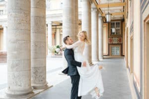 Ein Bräutigam im schwarzen Anzug hebt eine lächelnde Braut im weißen Kleid und Spitzentop mit einem Blumenstrauß hoch, während sie freudig unter hohen Säulen in einem elegant dekorierten Korridor posieren.