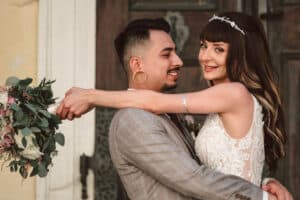 Braut und Bräutigam lächeln sich an, wunderschön eingefangen von Klickermann Photography. Der Bräutigam im karierten Anzug hält die Braut in ihrem weißen Spitzenkleid und ihrer Tiara fest. Sie umklammert einen Blumenstrauß, während die beiden liebevoll vor einer Holztür stehen.