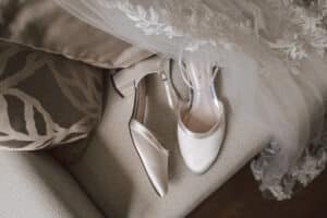 Ein Paar elegante elfenbeinfarbene Brautschuhe mit Blockabsatz stehen auf einem Sofa neben einem mit Spitze besetzten Brautschleier. Der Schleier ist mit aufwendigen Blumenstickereien verziert, und das Sofa hat gemusterte Kissen.