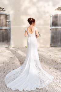 Eine Frau in einem weißen Spitzenbrautkleid steht draußen auf Kies und blickt von der Kamera ab. Ihr Haar ist hochgesteckt, und sie hält einen kleinen Blumenstrauß in der Hand. Im Hintergrund sind zwei Holztüren an einer cremefarbenen Wand zu sehen.