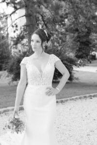 Eine Braut steht im Freien in einem weißen Brautkleid mit Spitzendetails, hält einen kleinen Blumenstrauß in der einen Hand und ruht mit der anderen Hand auf ihrer Hüfte, mit Bäumen und Grün im Hintergrund.